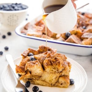 Slice of french toast casserole on a plate with blueberries being drizzled with maple syrup