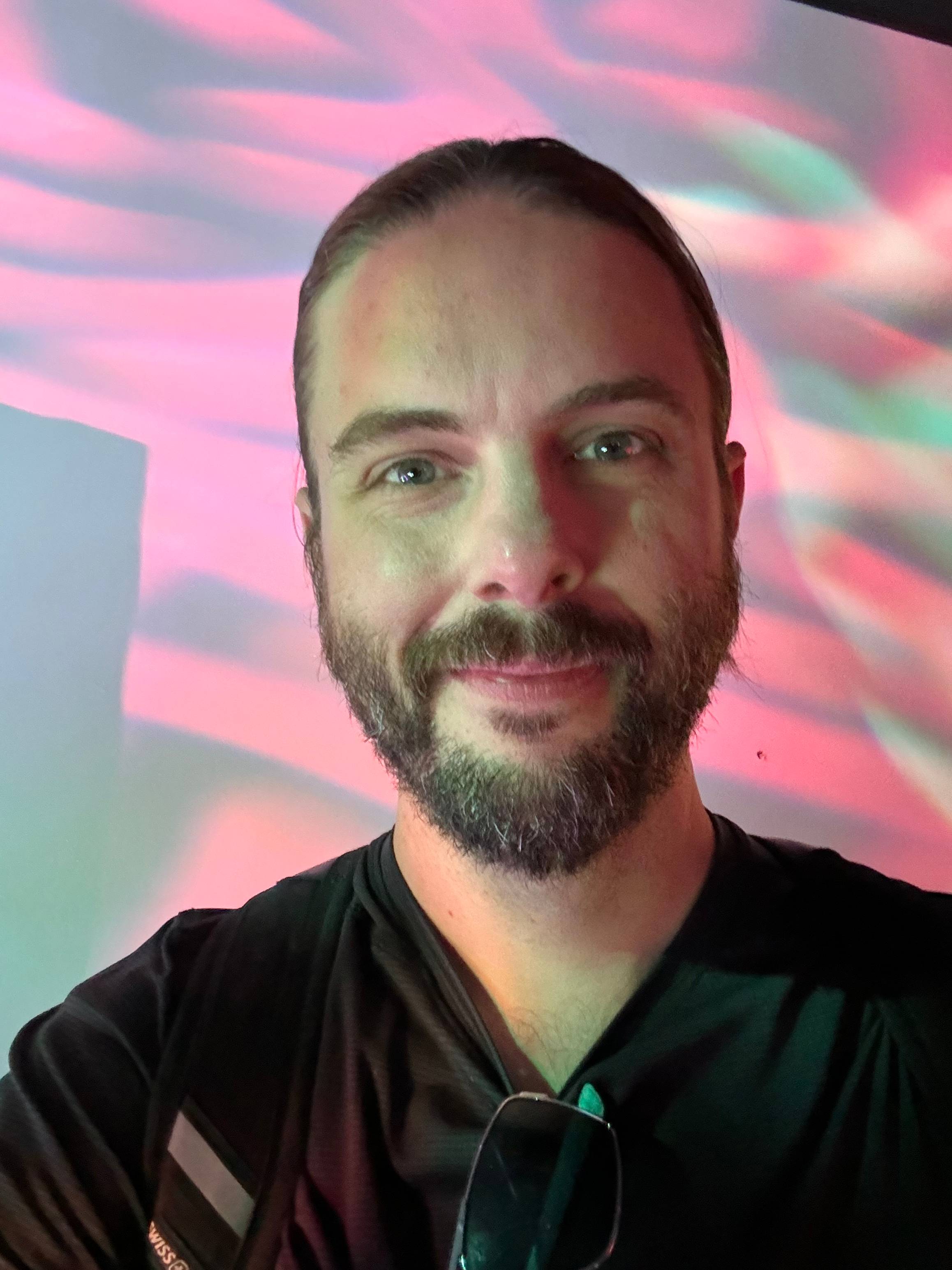 A person with long hair and a beard is smiling at the camera. They are wearing a black shirt, and there is a pair of sunglasses hanging from the neckline. The background features a colorful, wavy light pattern in shades of pink, red, and green.