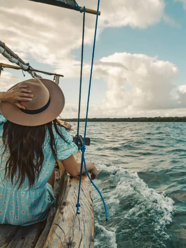 Woman with a blue dress and sun hat sitting in a boat in the middle of the ocean.
