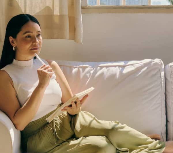 Woman with long black hair sitting on couch with pencil to her chin and notebook in hand.