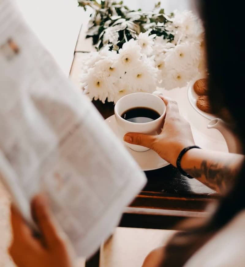 A person holding a coffee cup and reading a newspaper near white flowers