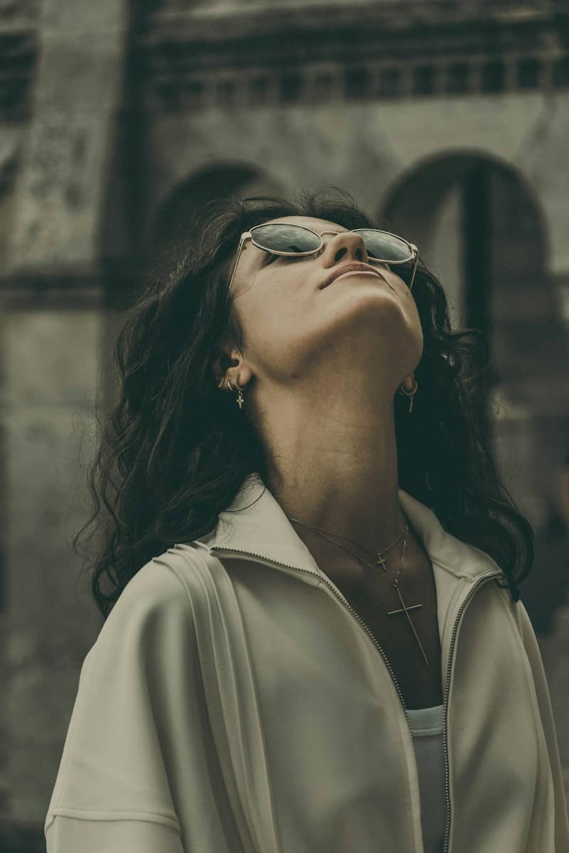 A woman with curly hair wearing sunglasses and cross necklaces looking upward