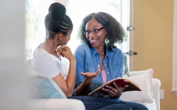 Two women sitting on a couch talking with an open bible in one of their hands.