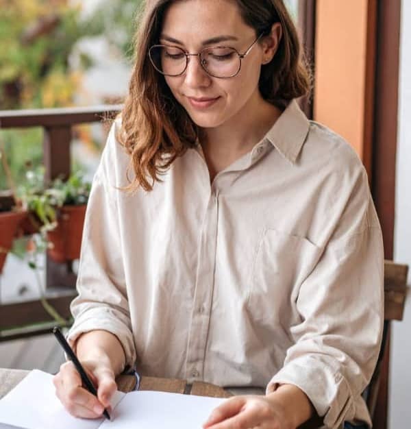 Woman with glasses siting at a table on the patio, writing in a journal.