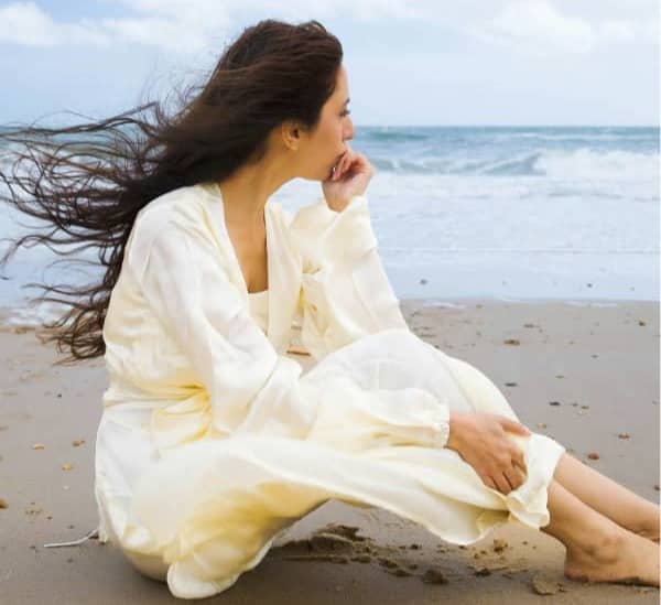 Mom wearing a cream dress, sitting on the sand near the shore of a beach.