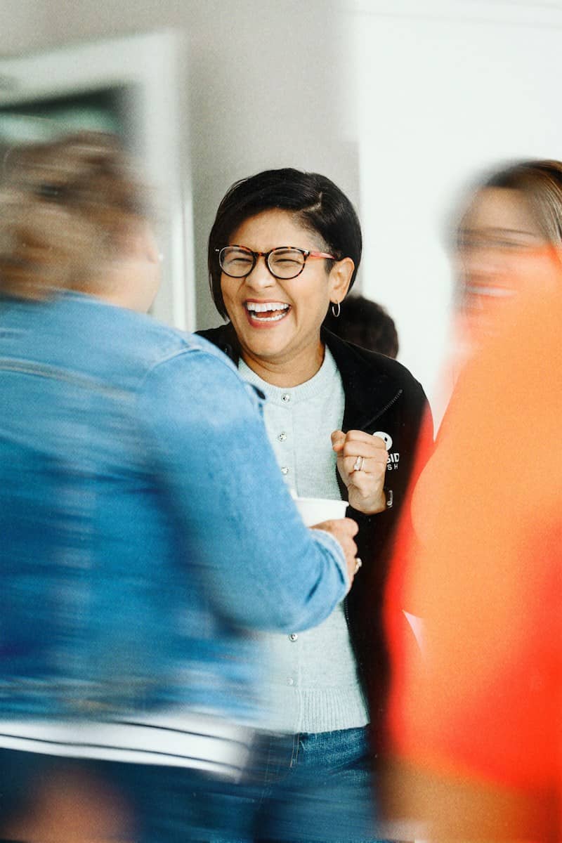 A woman with glasses laughing while talking to people in a blurred foreground