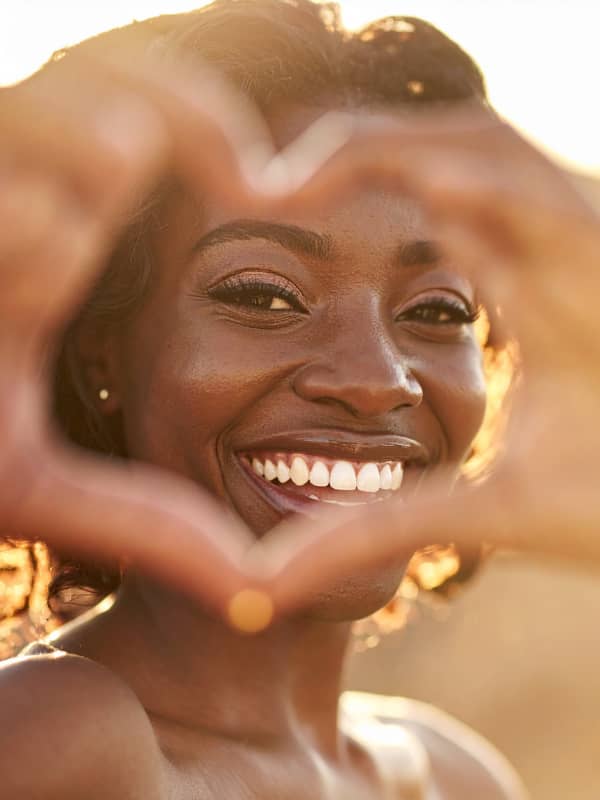 Portrait of a black woman smiling with heart hands in front of her face.