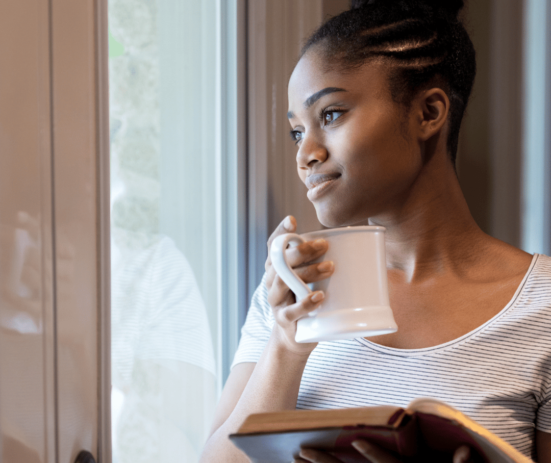 African American woman standing at a window with a coffee and book in her hand.