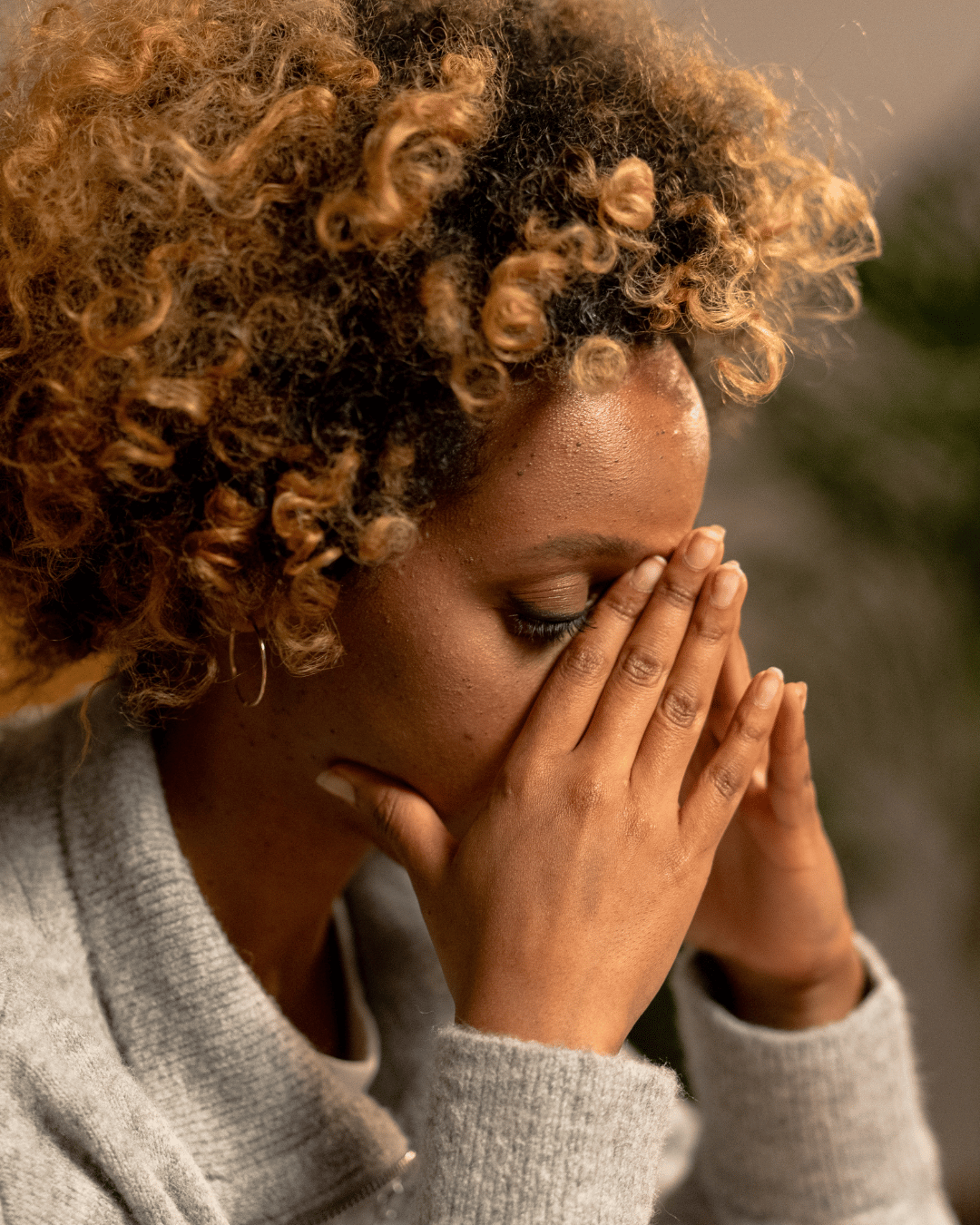African American woman with curls and gray sweater with face in her hands.