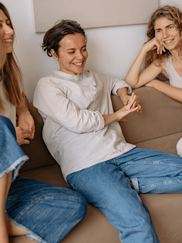 Three women sitting on a tan couch talking and smiling.