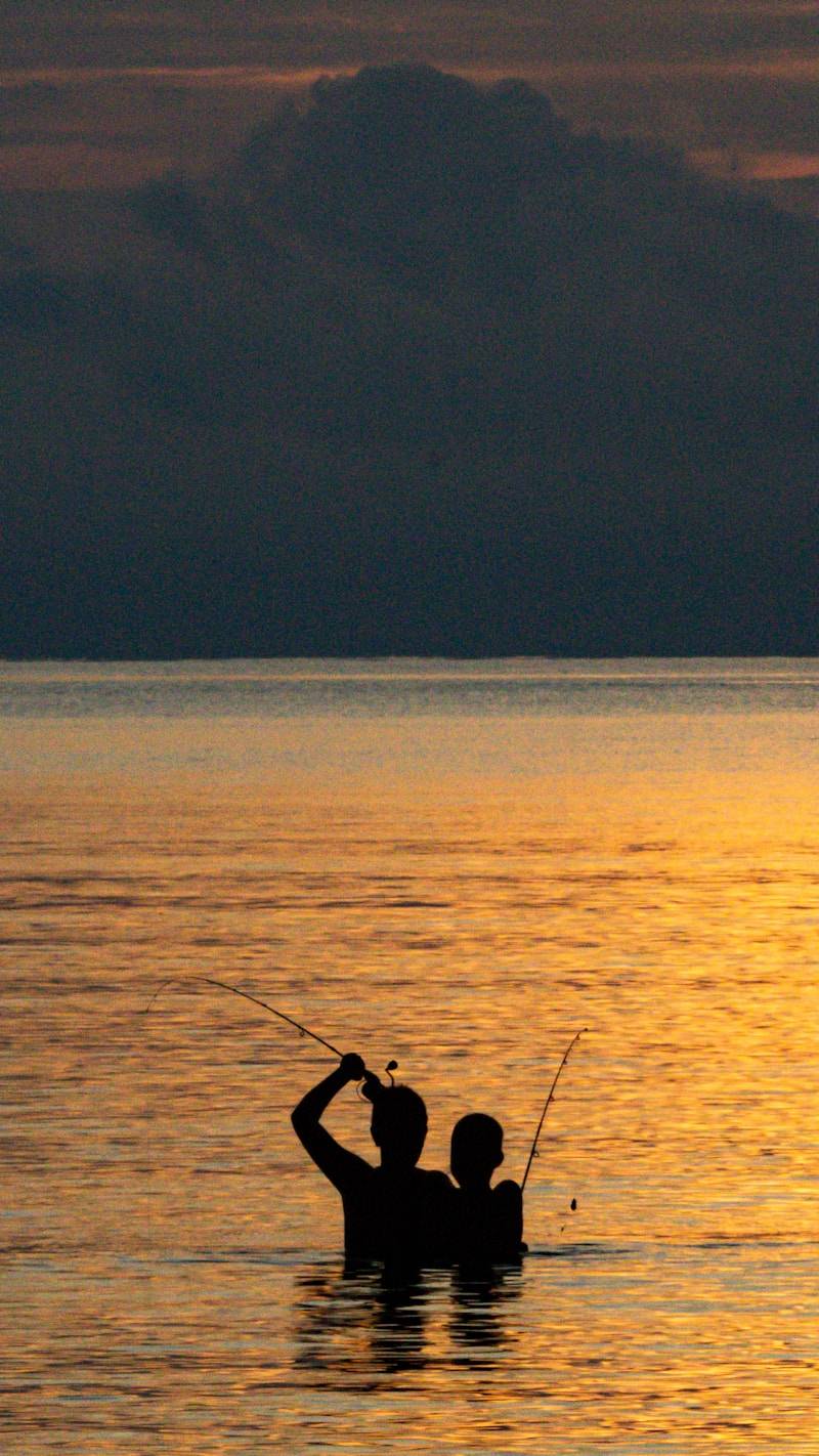 Two people fishing in the ocean at sunset.