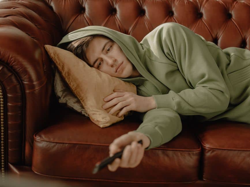 A young man in a green hoodie relaxes on a leather sofa with a remote control, depicting a cozy home scene.