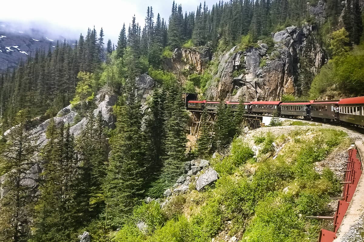 Train cars crossing a narrow trestle along a steep, forested mountainside on the White Pass Scenic Railway near Skagway, Alaska, surrounded by evergreen trees and rocky cliffs.