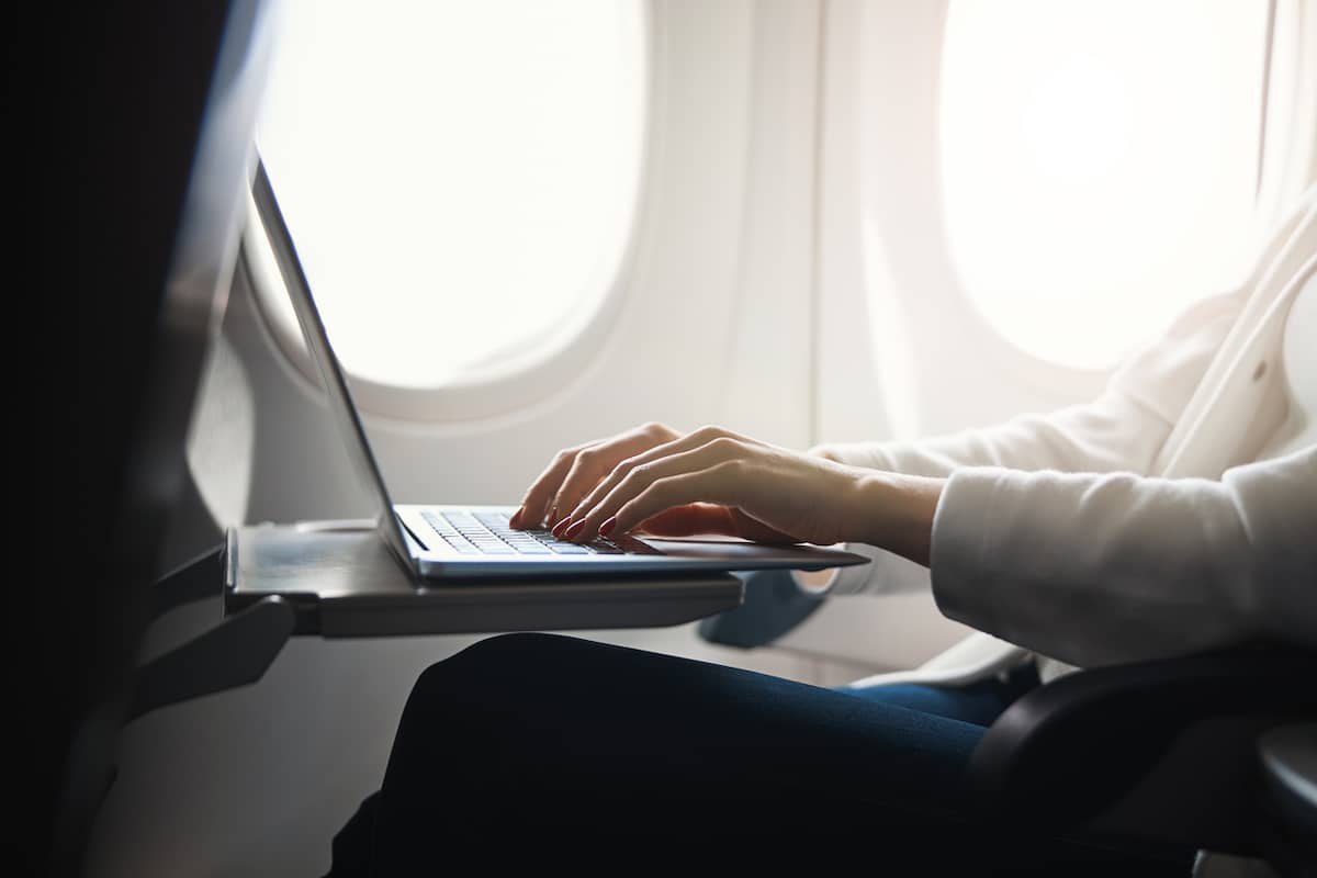 Close-up of a traveler's hands typing on a laptop at an airplane tray table beside a bright cabin window
