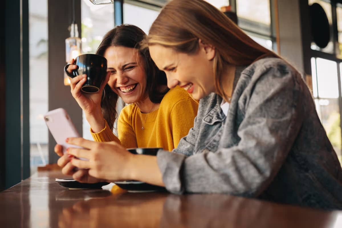 Two women laugh and smile as one holds a coffee mug and the other shows her phone so they moth can see it