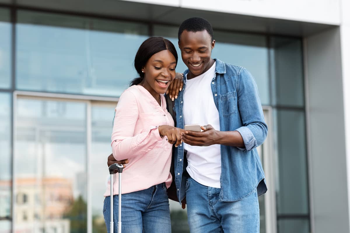 Couple standing outside an airport with luggage looking at a smartphone together.