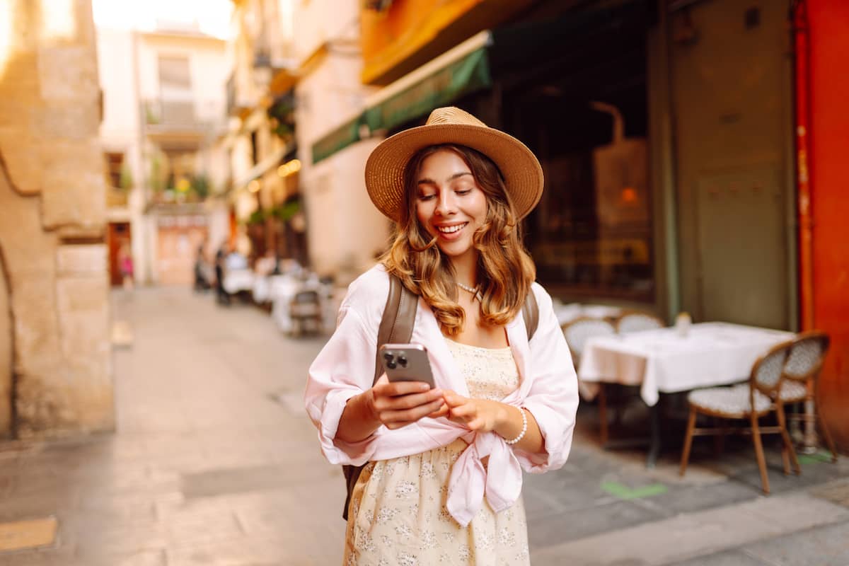 Young woman in a sun hat walks down a sunlit European street holding her phone, with outdoor café seating and stone buildings in the background.