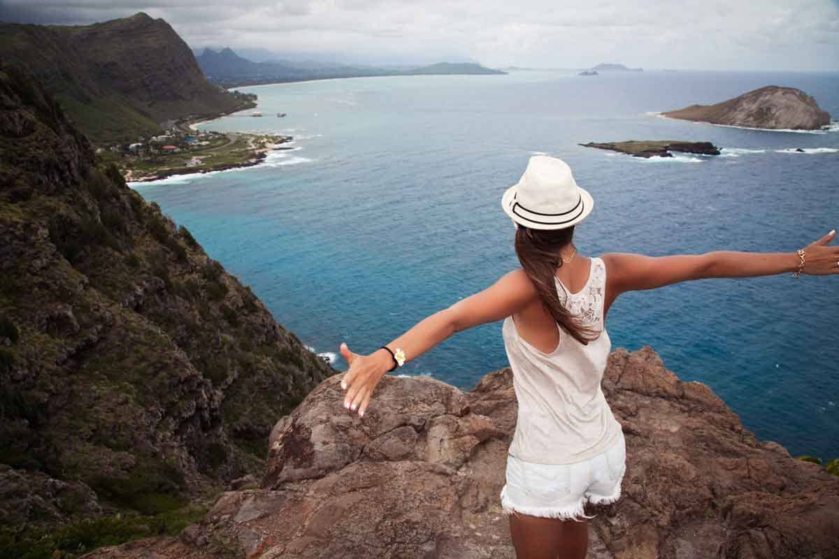 A female tourist standing on a rock with raised hands and looking down at a at view of beautiful Hawaiian landscape.