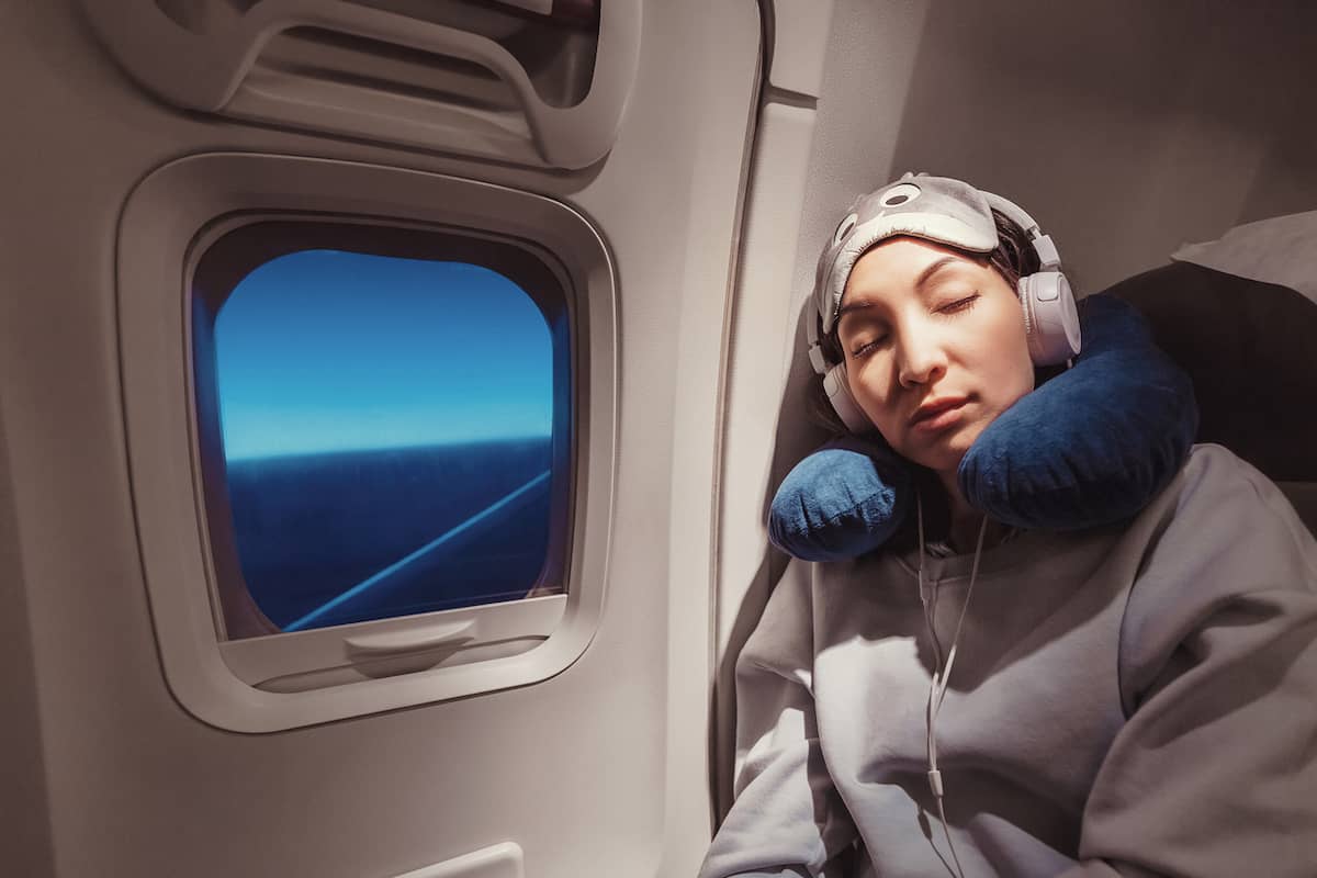 Woman sleeping against an airplane window with a neck pillow, eye mask pushed up on her forehead, and over-ear headphones. 