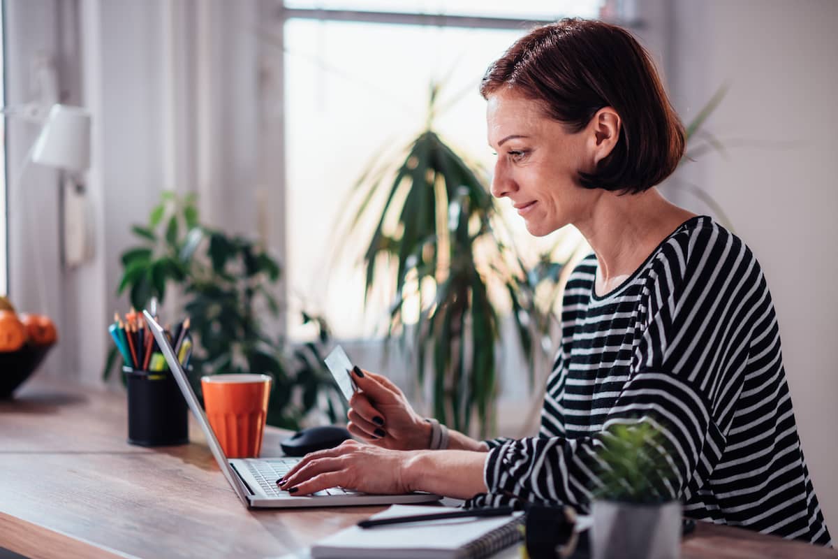 Woman with short dark hair sitting at a desk, looking at her laptop screen while holding a credit card in one hand.
