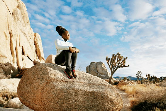 A woman wearing athletic clothing sits on a large rock in the open air of a dry and arid national park in the United States