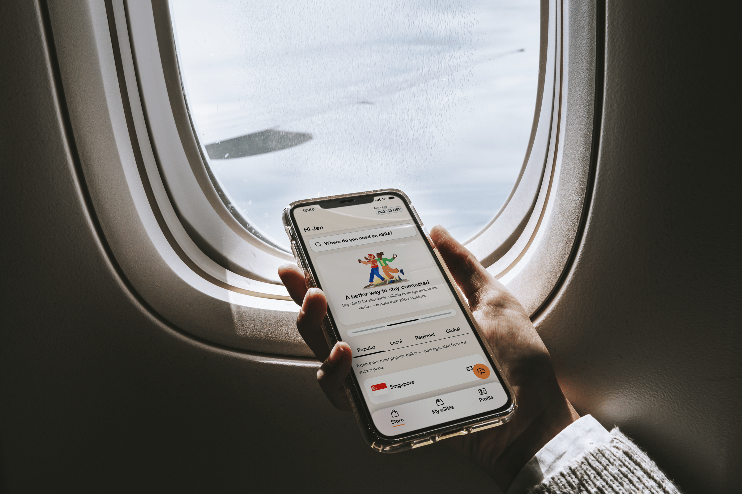 A hand in front of an airplane window in flight holds a mobile phone with the Airalo app shown on the screen.