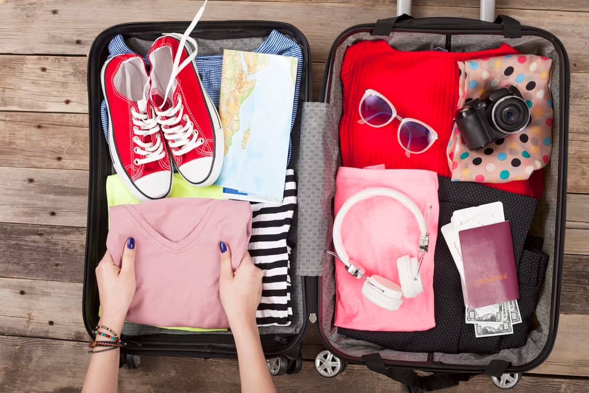 Hands packing a carry-on suitcase with clothing, red sneakers, headphones, sunglasses, a map, a passport, and a camera