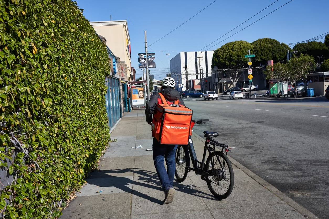 A DoorDash courier walks a bicycle down a sunny sidewalk, wearing an orange delivery backpack.