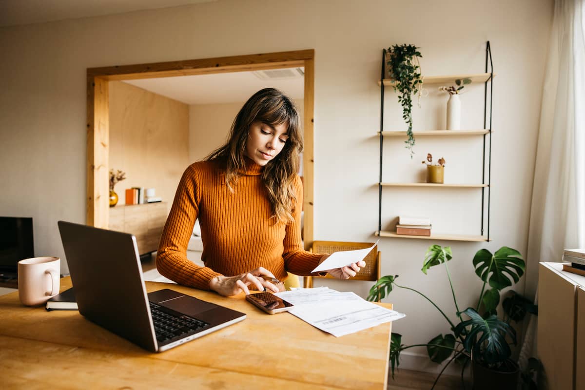 Woman in an orange sweater using a calculator and reviewing bills at her desk, with a laptop and houseplants nearby.