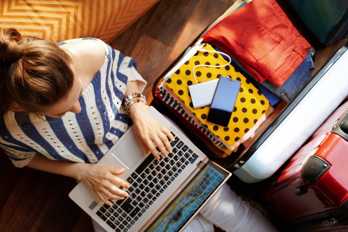 Traveler using a laptop while packing a suitcase for a trip, with clothes and travel accessories visible.