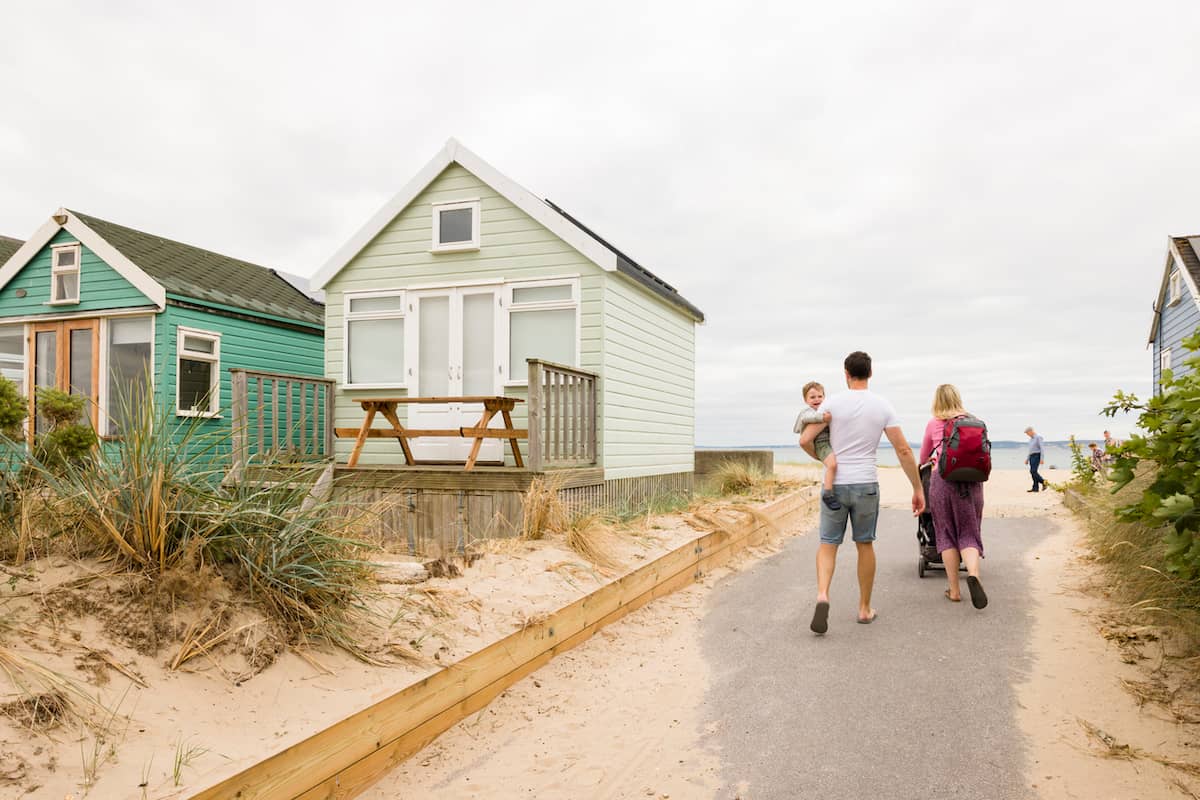 Family walks past pastel beach huts toward the shore on a summer holiday in Dorset, U.K.