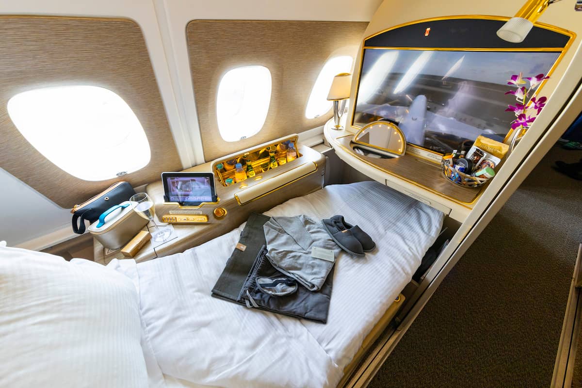Emirates Airbus A380 first-class suite with lie-flat bed by the window, large entertainment screen, minibar, amenity kit, and pajamas laid out on the bed.