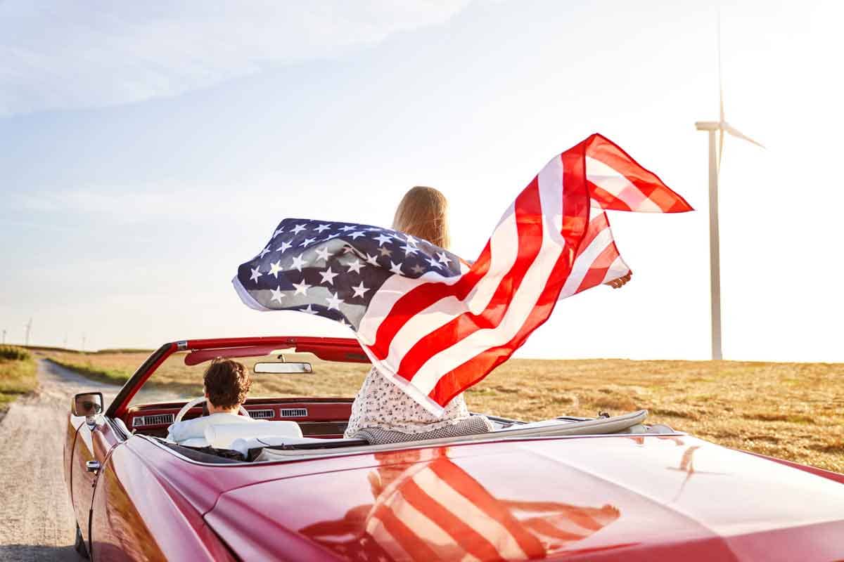 A woman holds an American flag standing on the seat of a convertible on a road trip.