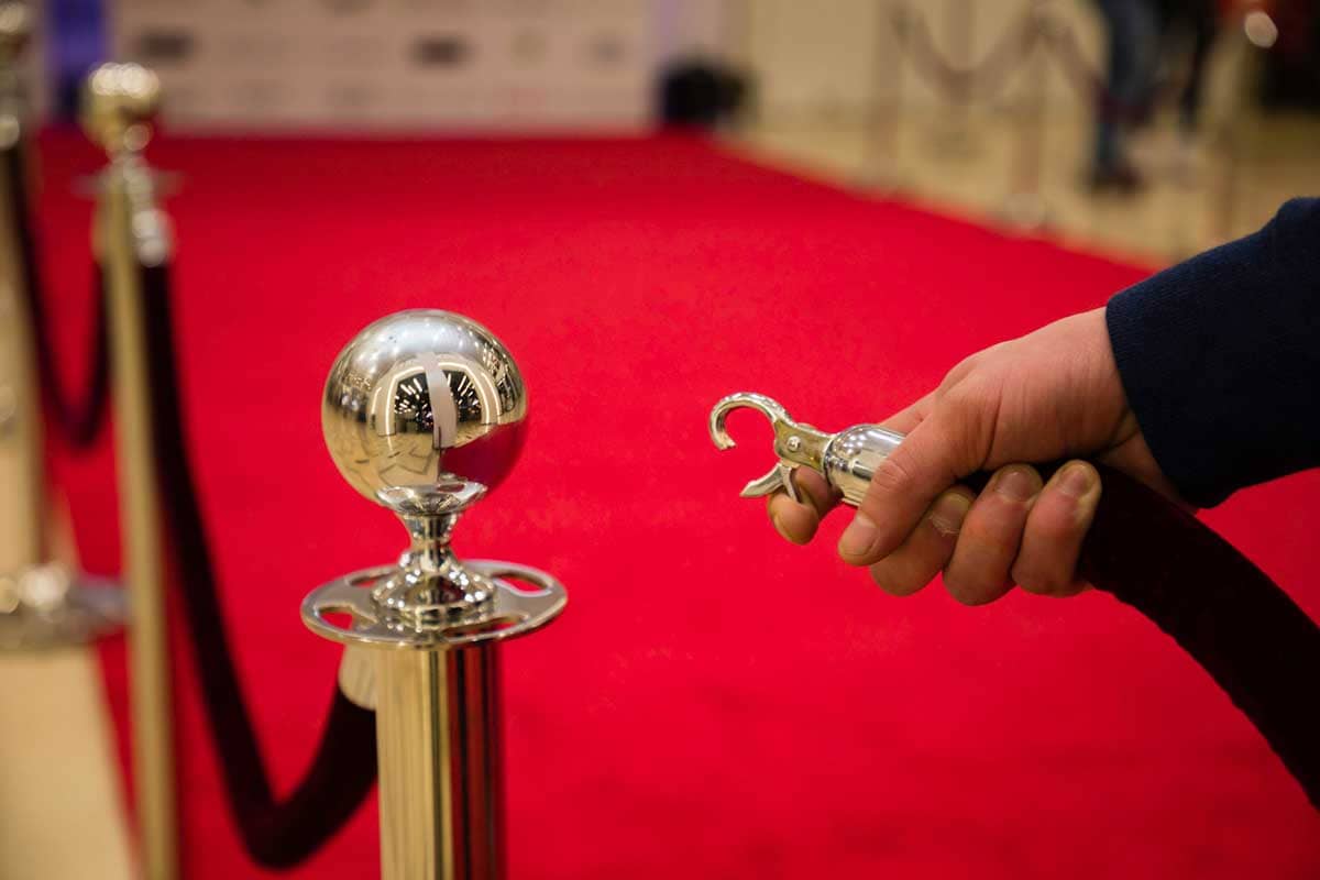 A hand opens a velvet rope to a red carpet