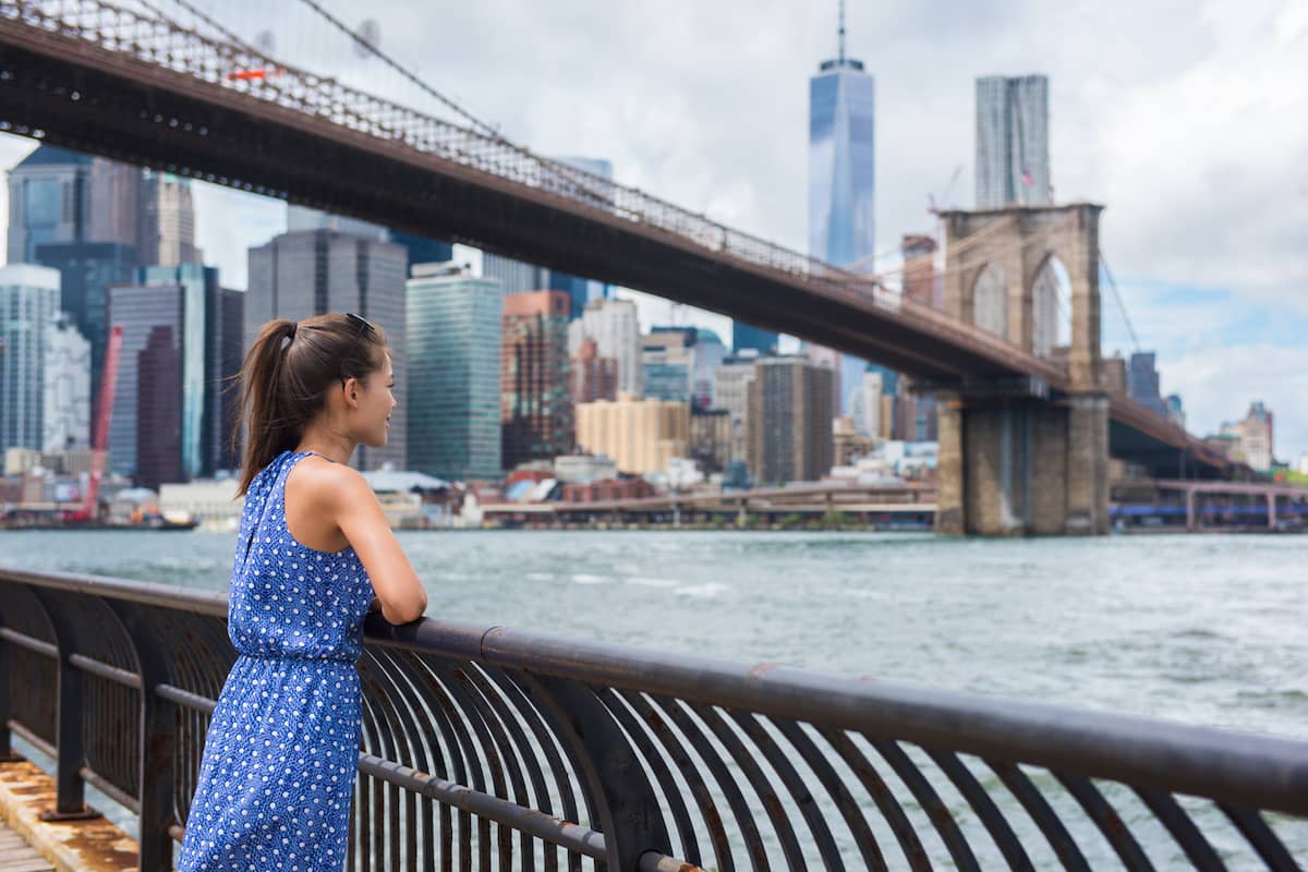 Woman at a waterfront railing taking in the Brooklyn Bridge and Manhattan skyline.