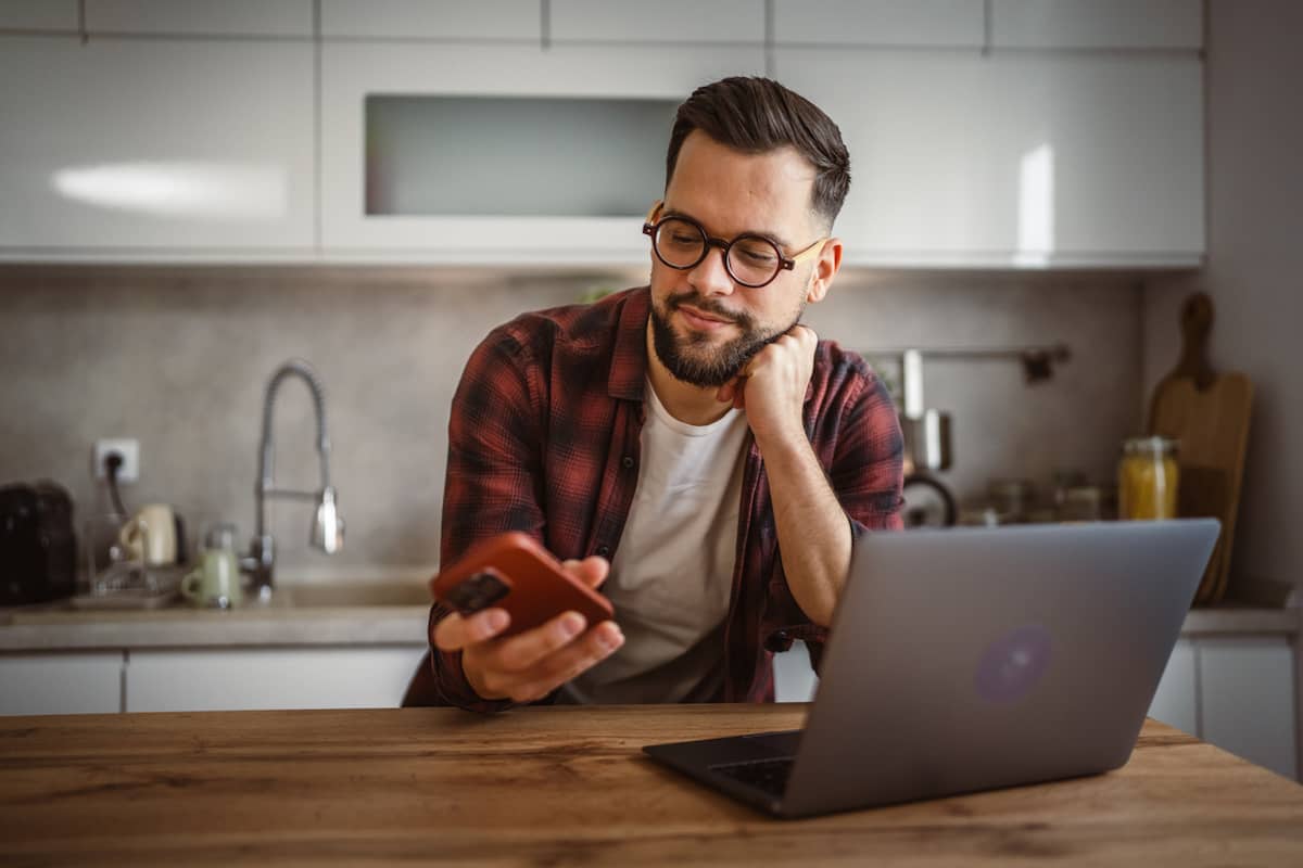 A man leans on his kitchen counter and reads a notification on his phone next to an open laptop. 