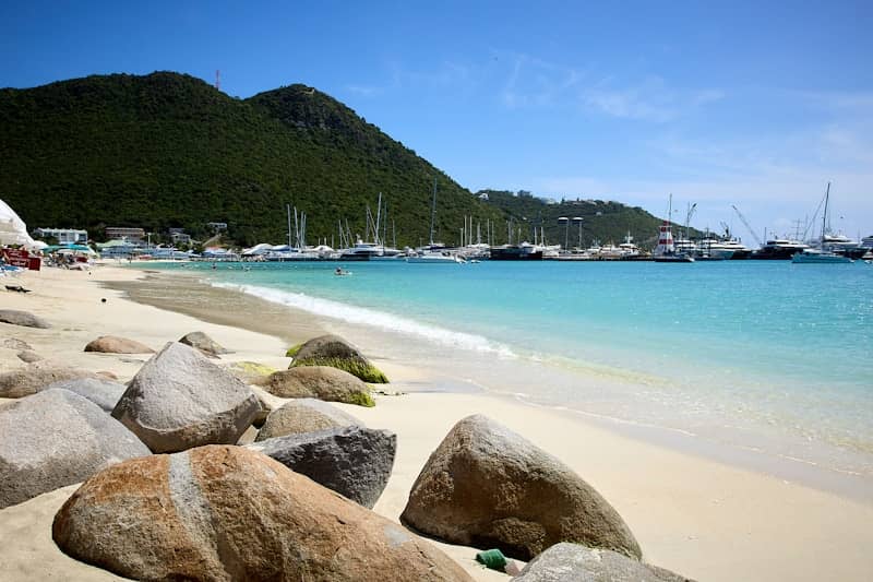 Rocks on a sandy beach with turquoise water and boats.