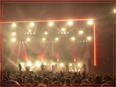 Concert venue with a packed crowd facing a brightly lit stage framed by warm amber and white lights.