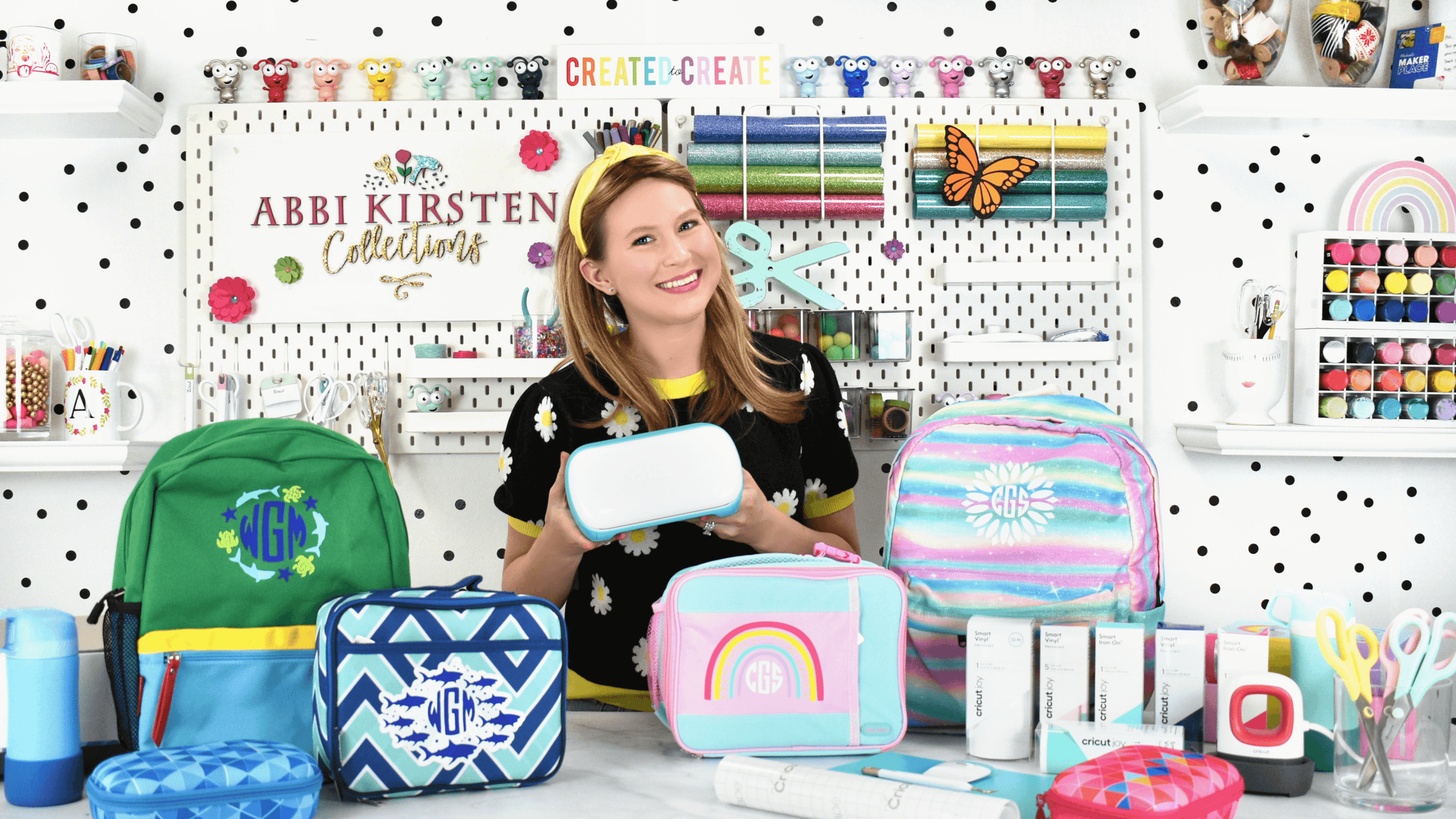 The image shows Abbi Kirsten surrounded by lunch boxes and backpacks that have been customized with iron-on vinyl. 