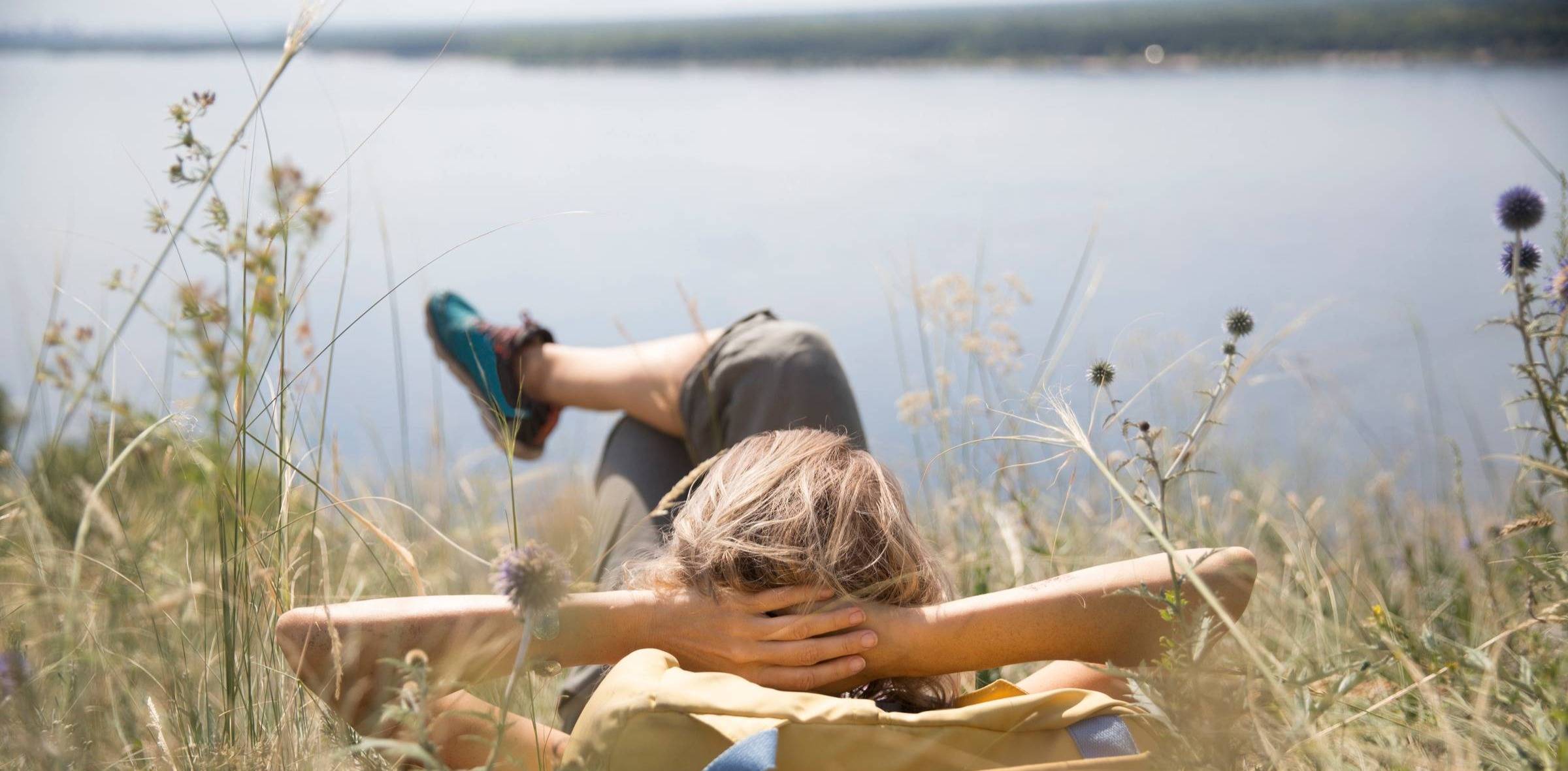 woman lying on dry grass with hands behind head looking out over a lake