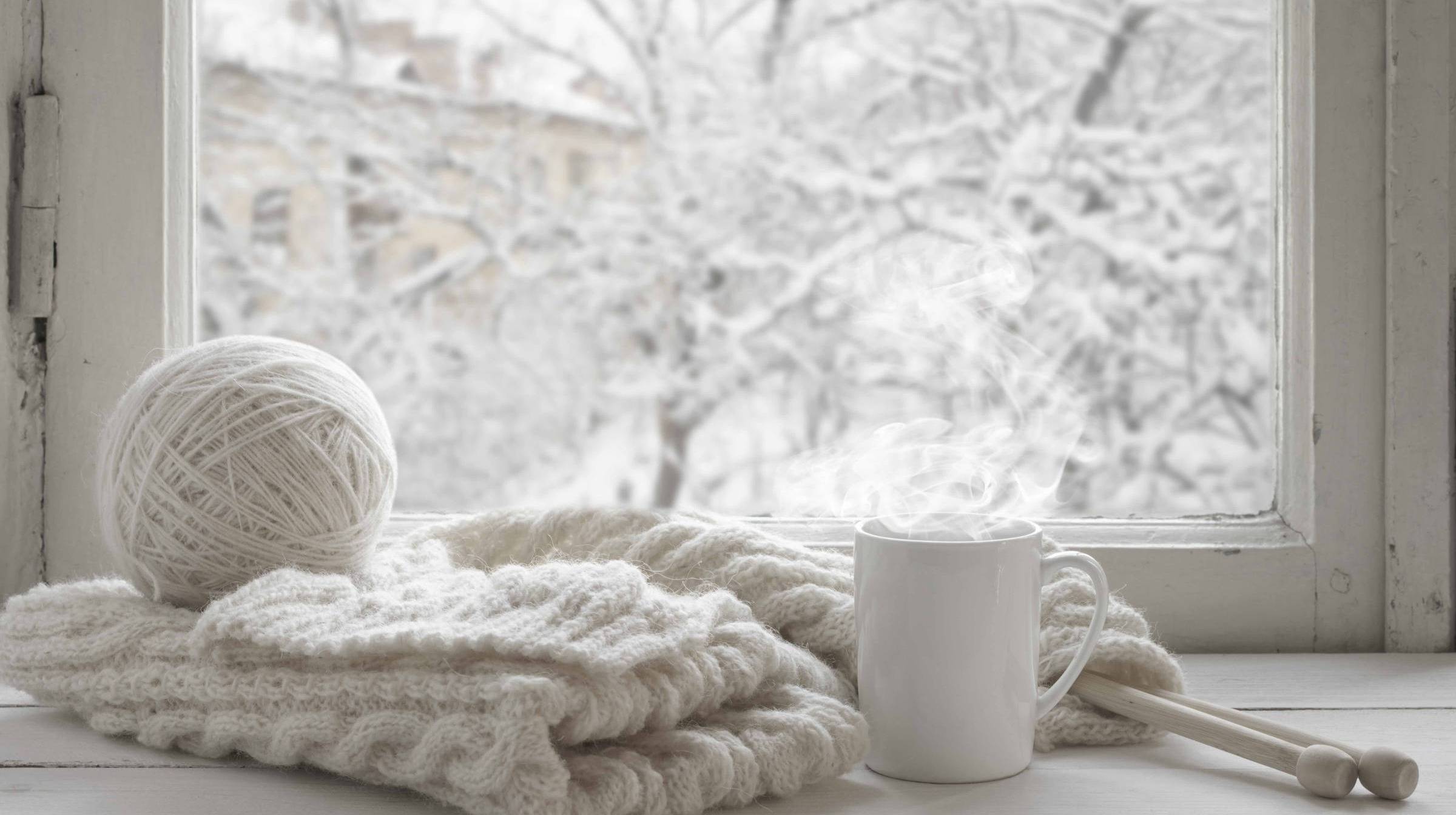 white winter scene of ball of white yarn and knitting, with a white mug and snow covered trees in the background