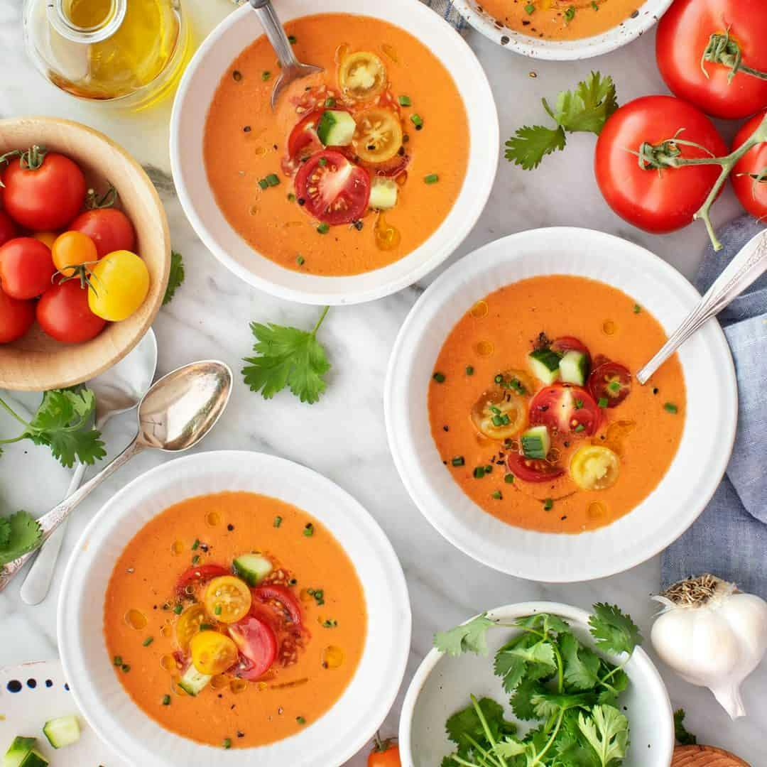 Three bowls of gazpacho topped with cherry tomatoes, cucumber, and chives
