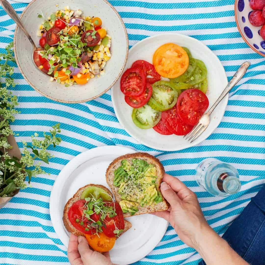 Overhead shot of picnic scene with tomato and corn salad, plate of sliced tomatoes, and hands assembling tomato and avocado sandwich on a plate