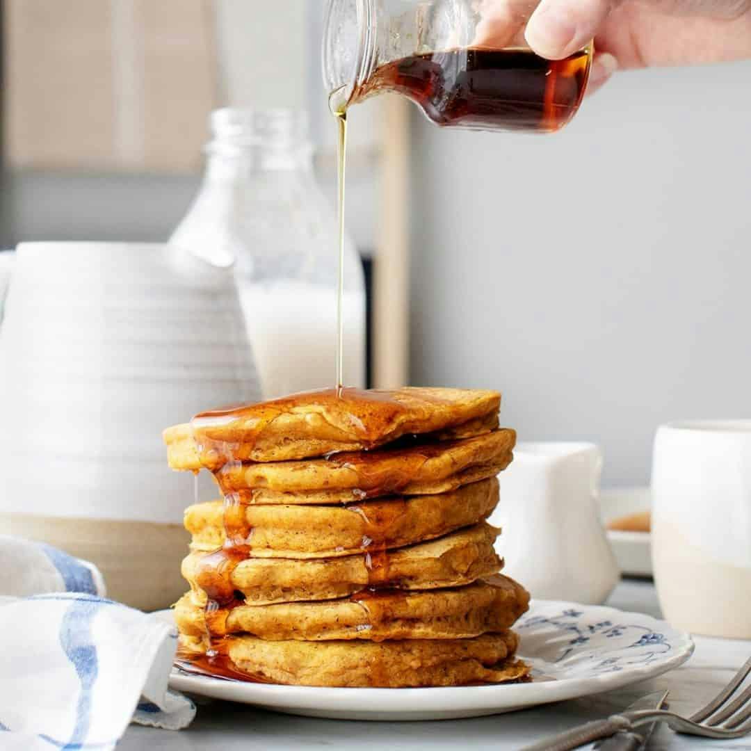 Hand pouring maple syrup over stack of pumpkin pancakes