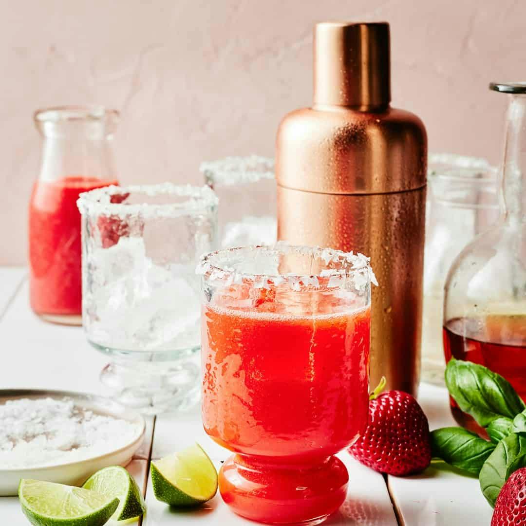 Strawberry margarita with cocktail glasses, cocktail shaker, and lime wedges