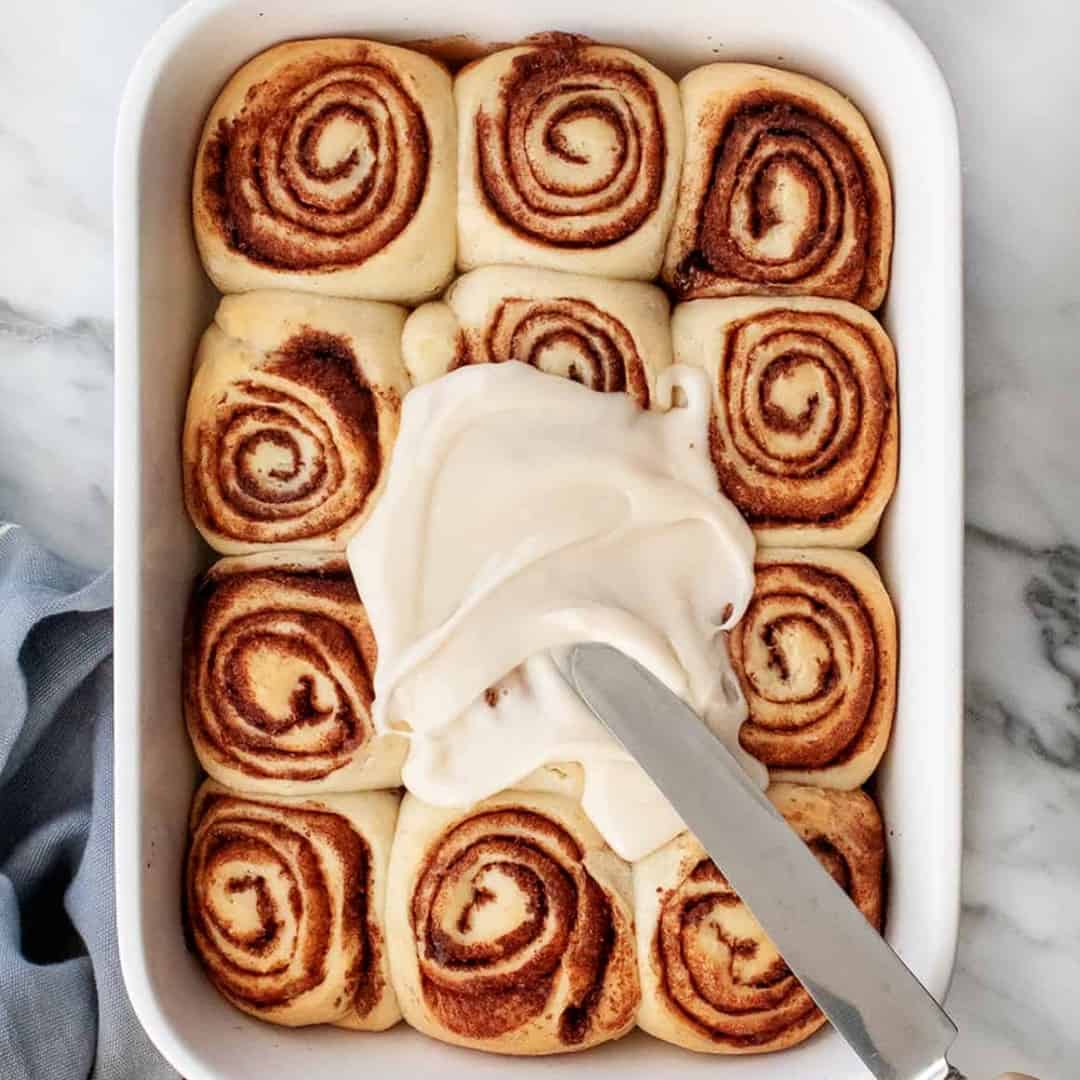 Knife spreading glaze on cinnamon rolls in baking dish 