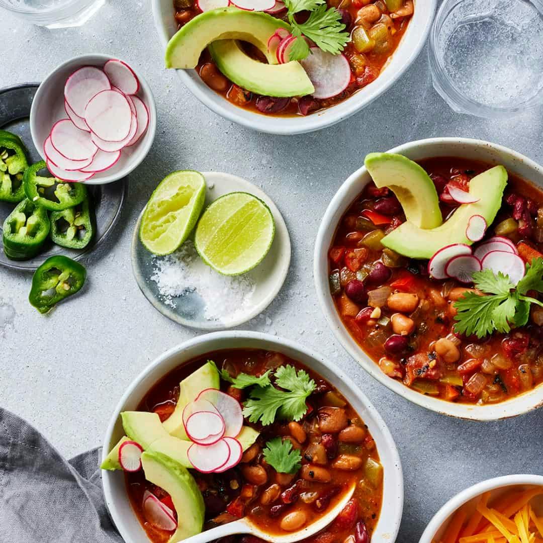 Three bowls of chili topped with avocado slices, radishes, and cilantro