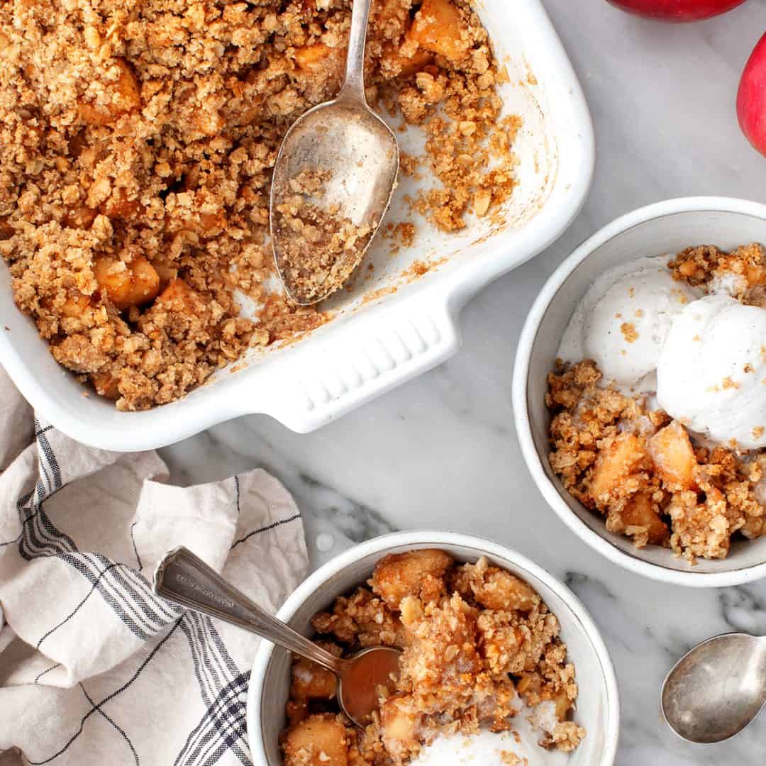 Apple crumble in baking dish next to two servings in bowls with vanilla ice cream