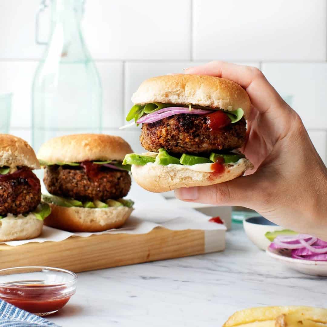 Hand holding veggie burger on a bun with red onion, avocado, lettuce, mayo, and sriracha