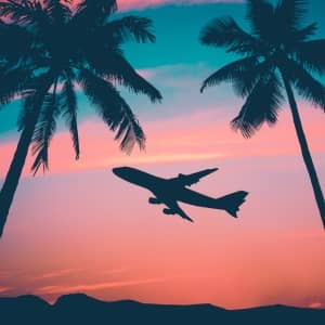 A plane flying to a tropical location in a sunset of orange, pink and teal colors.